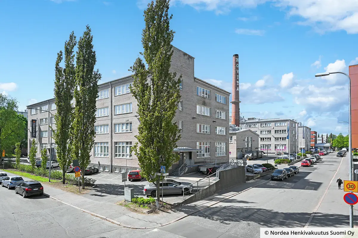 Toimitilakiinteistö Helsingin Kiviaidankatu 2 kuvattuna Vattuniemenkadun kulmauksesta. Talon pihassa punatiilinen vanha savupiippu. Talon edessä pysäköityjä autoja ja puita.