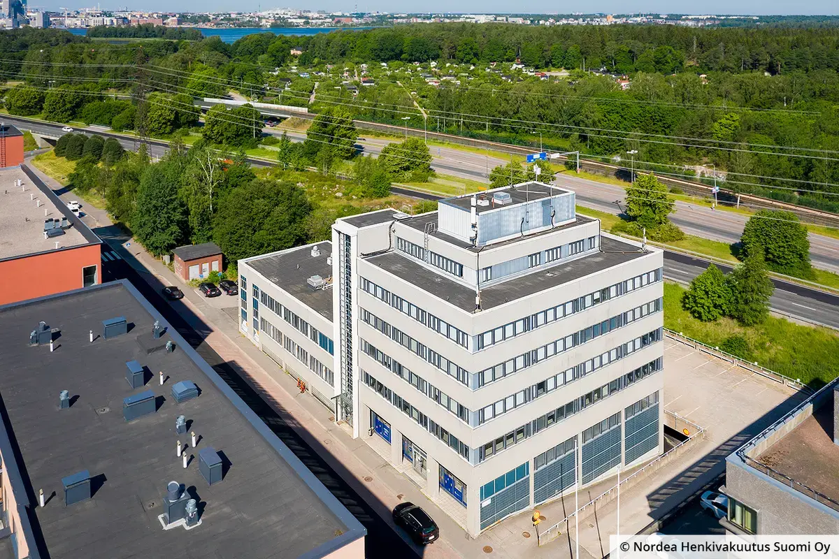 Helsingin Hitsaajankatu 4 -toimitilakiinteistö ilmakuvassa. Harmaa teollishenkinen rakennus on Itäväylän varrella, pihassa pysäköintitilaa kahdessa tasossa.