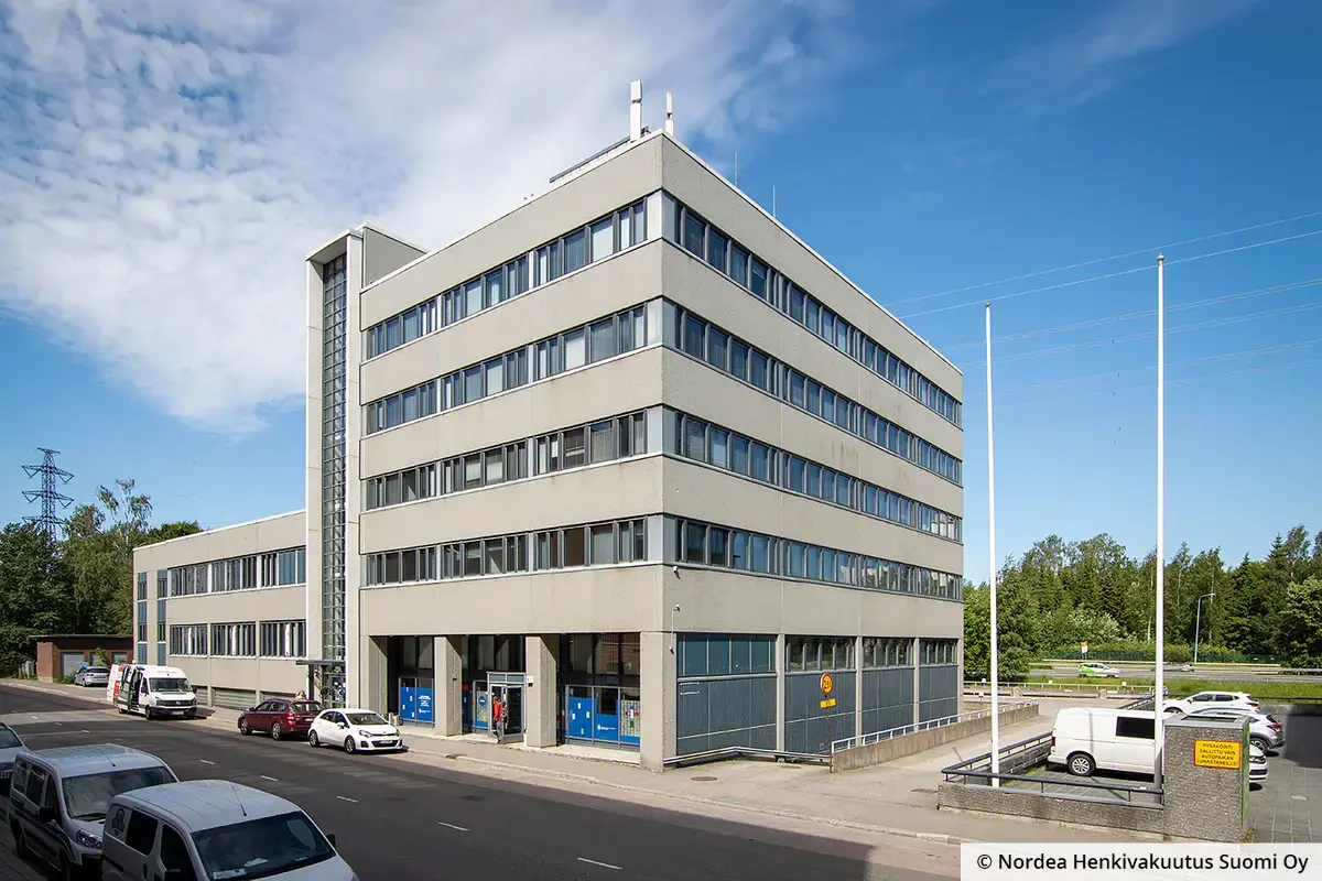 Helsingin Hitsaajankatu 4 harmaa julkisivu. Korkeammasta ja matalammasta osasta muodostuvan kiinteistön kerroksissa on nauhaikkunat, katutasossa liiketilaa.