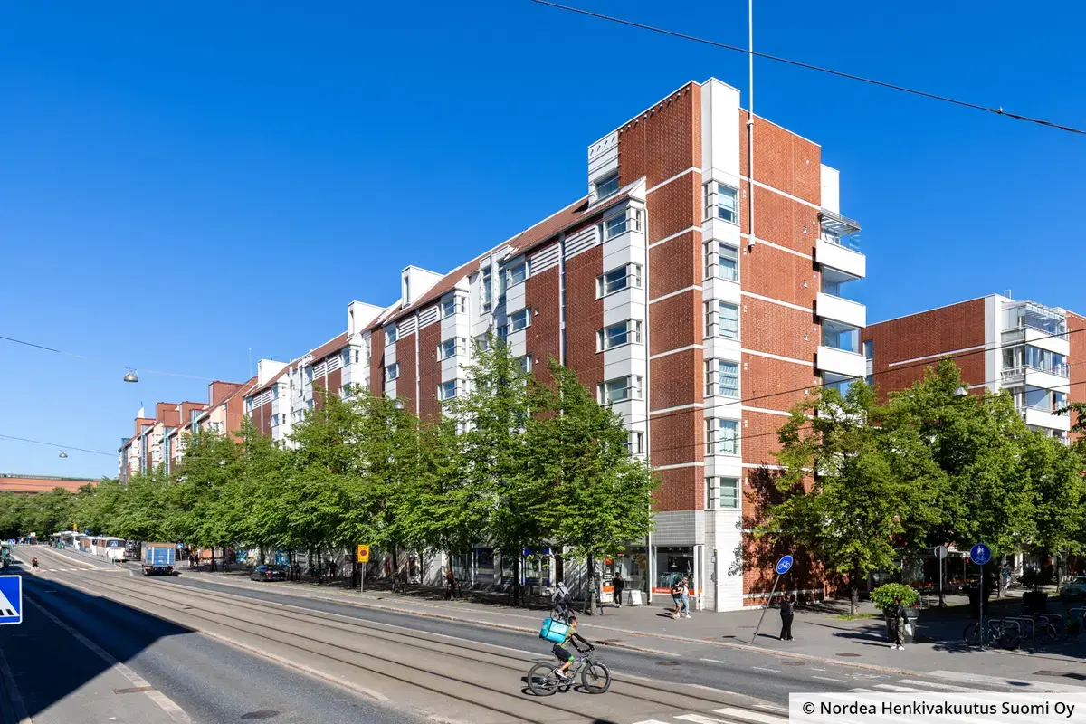 Kiinteistön julkisivu on vaalea ja punatiilinen, katutasossa on liiketiloja. Talon edessä kulkee autotie ja ratikkakiskot.
