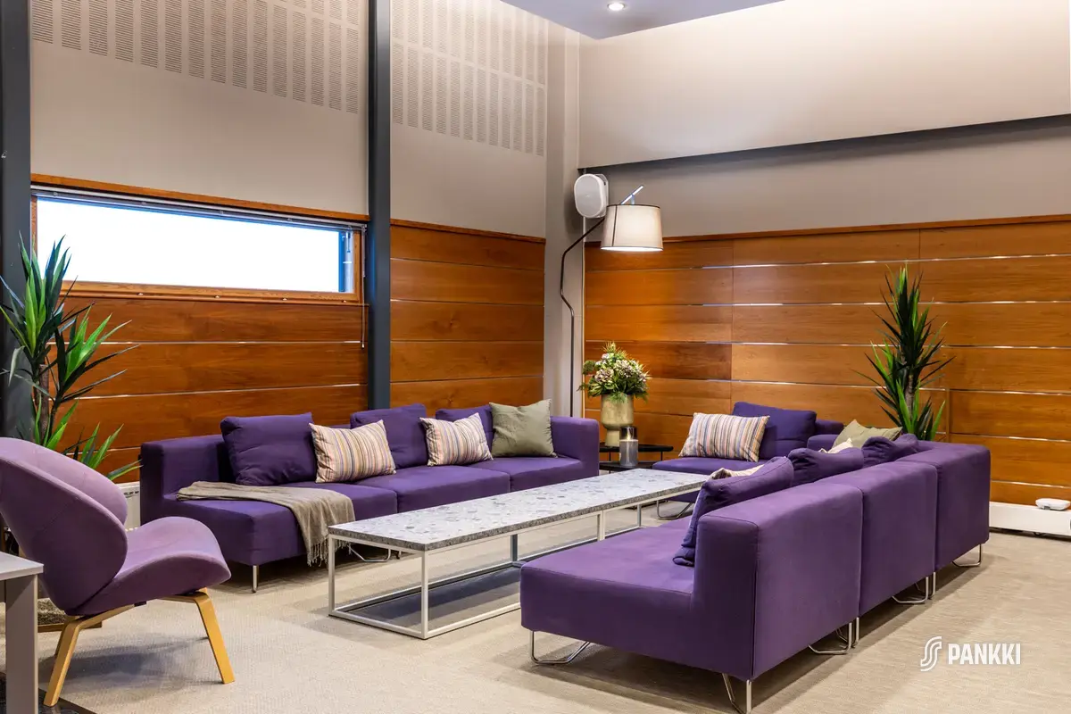 Toimistotalon saunaosaston lounge-tilassa on violetti sohvaryhmä ja marmorikantinen pitkä sohvapöytä. Seinät ovat tumman ruskeat. Kolme viherkasvia on taustalla.