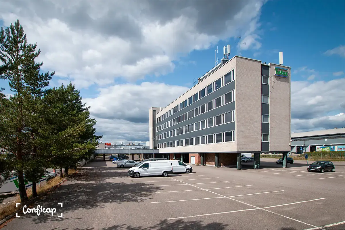 Toimitilakiinteistön Vantaan Tikkurilantie 10 toimistotorni ja etelänpuolimmainen tuotantotilasiipi kuvattuna pysäköintialueelta. Toimistotalon ruskea julkisivu kylpee auringossa. Pihalla autoja.