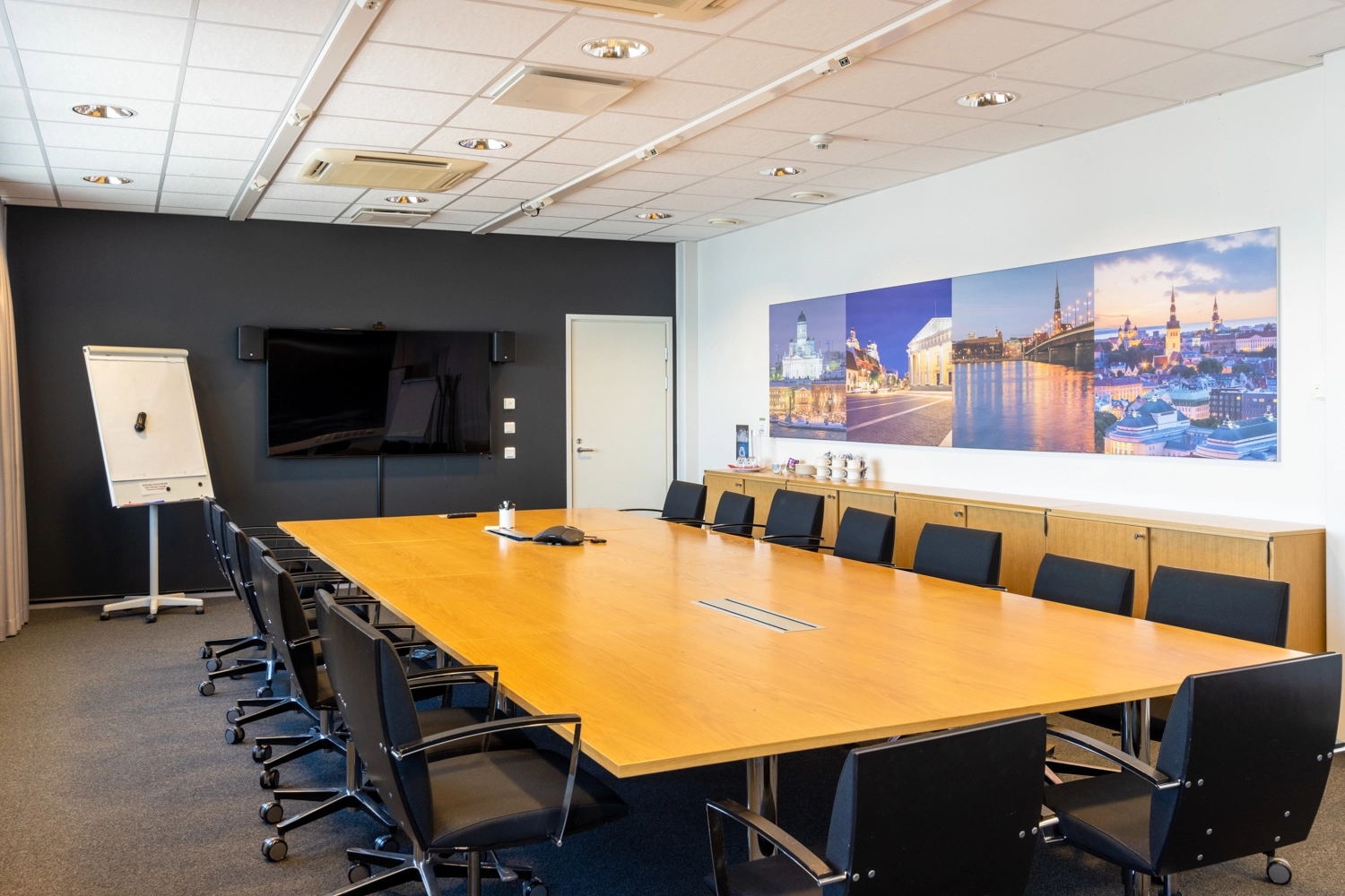 Toimiston neuvotteluhuoneessa on iso kokouspu00f6ytu00e4 ja 15 tuolia sen ympu00e4rillu00e4. Screen on kiinnitetty seinu00e4lle.
