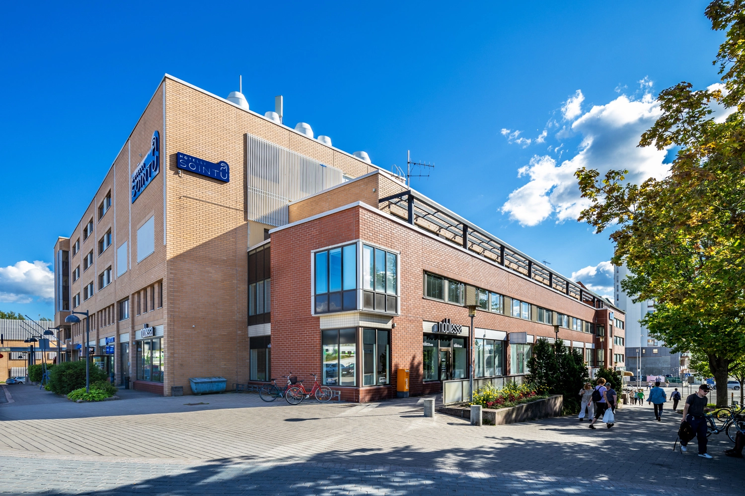 Ju00e4rvenpu00e4u00e4n Asemakatu 4:n julkisivu Kaksivu00e4risen kiinteistu00f6n seinu00e4ssu00e4 on hotellin ja katutason liiketilojen kyltit, edessu00e4 pyu00f6ru00e4teline ja paljon ku00e4veliju00f6itu00e4.