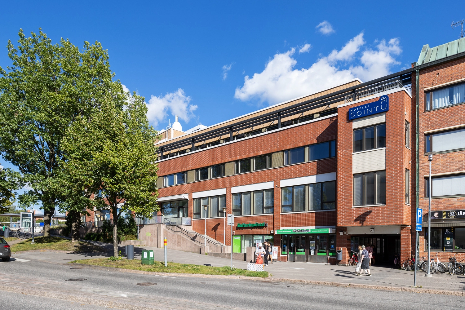 Ju00e4rvenpu00e4u00e4n Asemakatu 4 punatiilisen siiven julkisivu. Katutasossa liiketila, jonka edessu00e4 jalankulkijoita.