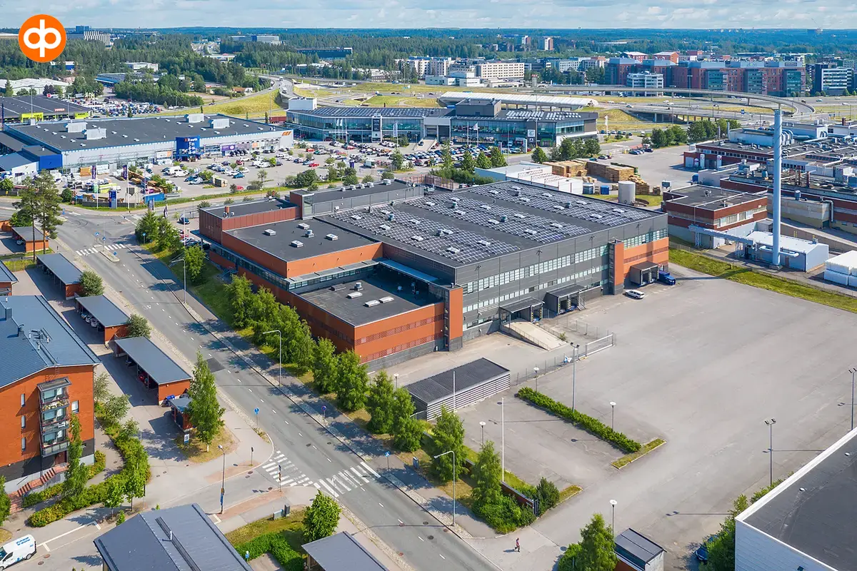 Red-gray commercial property Silvastintie 1 in Vantaa, aerial view from the south. There is an asphalted yard in front of the building. In the background are other commercial buildings and the airport