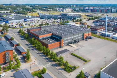 Red-gray commercial property Silvastintie 1 in Vantaa, aerial view from the south. There is an asphalted yard in front of the building. In the background are other commercial buildings and the airport