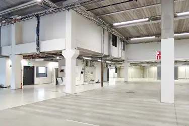 A light-colored warehouse in Vantaa. The space has a couple of supporting pillars, and there are building services on the roof. On the left is a so-called small mezzanine floor with office space.