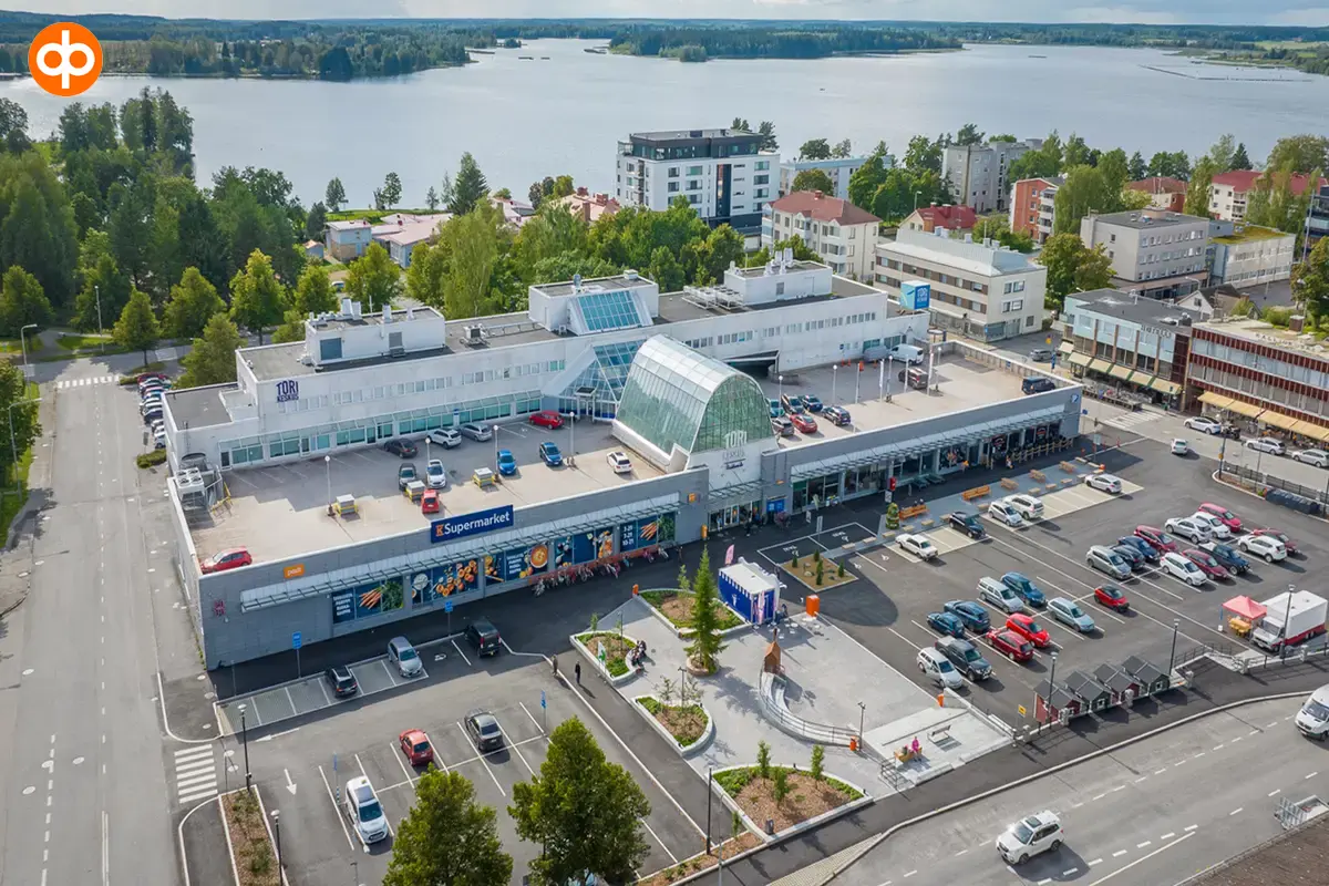 Aerial view of Sastamalan Torikeskus. Shopping centre with a large parking area in front of it. In the background residential buildings and a body of water. 
