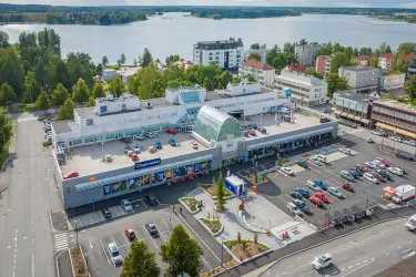 Aerial view of Sastamalan Torikeskus. Shopping centre with a large parking area in front of it. In the background residential buildings and a body of water. 