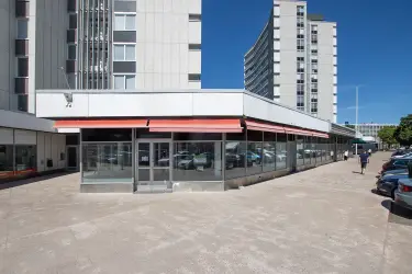 Kuva katutason liiketiloista asuinrakennuksen katutasossa. Liiketilassa on isot ikkunapinnat ja kiinteistön edessä on reilusti parkkipaikkoja.