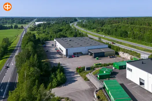 Järvenpää Tempo 4, aerial view of the business premises. A light, box-shaped building with a flat roof and an asphalted yard around it. Surrounded by trees and fields.