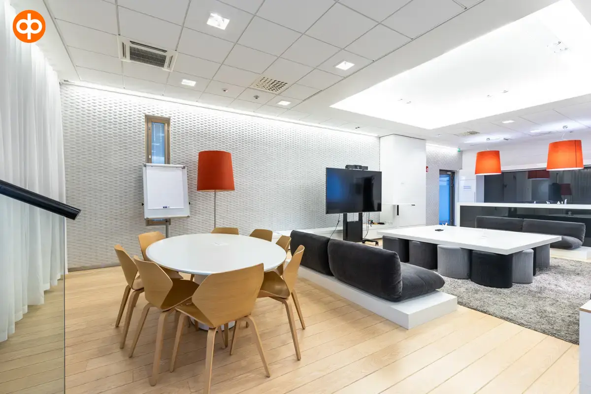Alberga Business Park, the meeting room of the office property. In the middle of the room, a group of tables and a sofa area, in the space three lamps with red lampshades. A kitchen in the background.