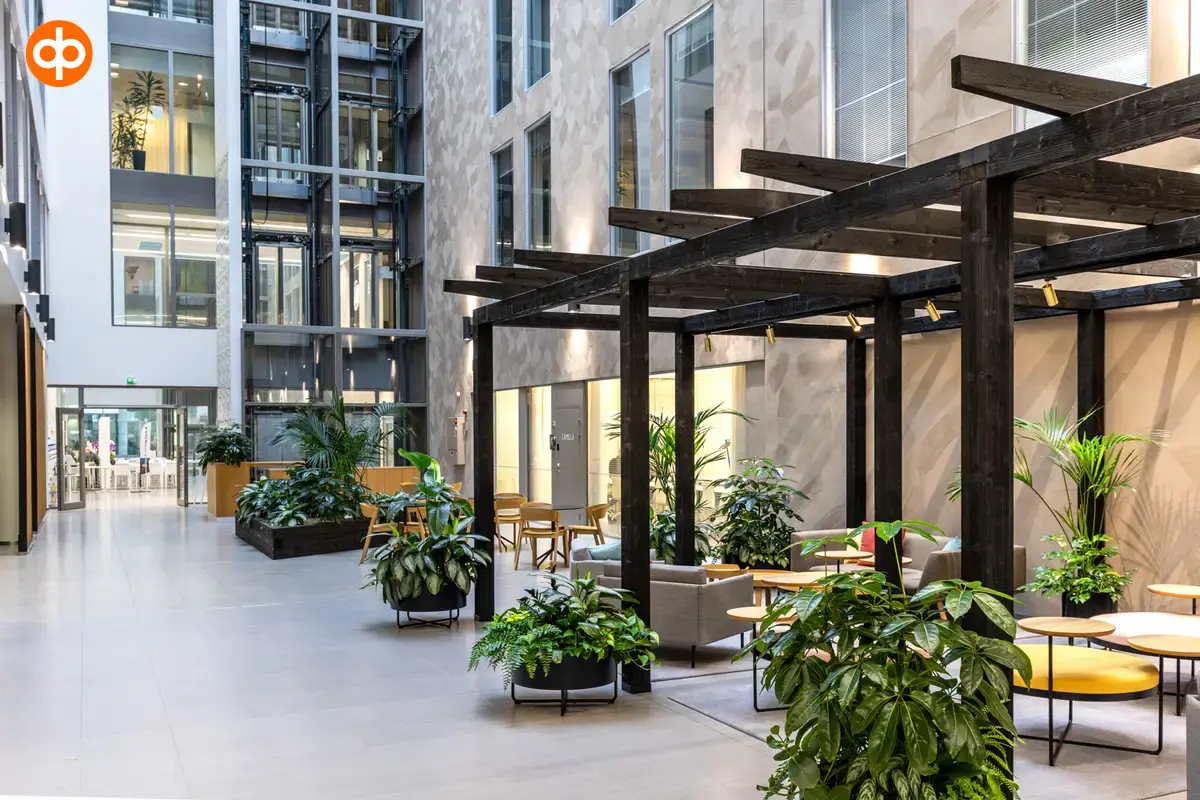 Alberga Business Park, a high-ceilinged lounge area with windows to office spaces above. In the lounge, groups of sofas surrounded by large green plants.
