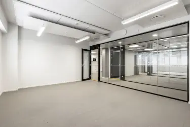 Helsingin Lönnrotinkatu 11, toimitilan sisäkuva. Tyhjä toimistohuone, jossa harmaat lattiat ja vaaleat seinät. Käytävän suuntaan oleva seinä lasia, karmit ja ovi mustat. 