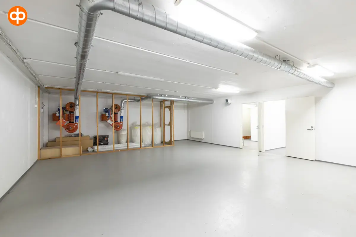 Interior view of the Vantaa Silvastintie 1 office building. Empty space with a mesh cage on the back wall, with stuff inside, light surfaces, large metal pipes on the ceiling.