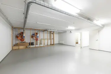 Interior view of the Vantaa Silvastintie 1 office building. Empty space with a mesh cage on the back wall, with stuff inside, light surfaces, large metal pipes on the ceiling.
