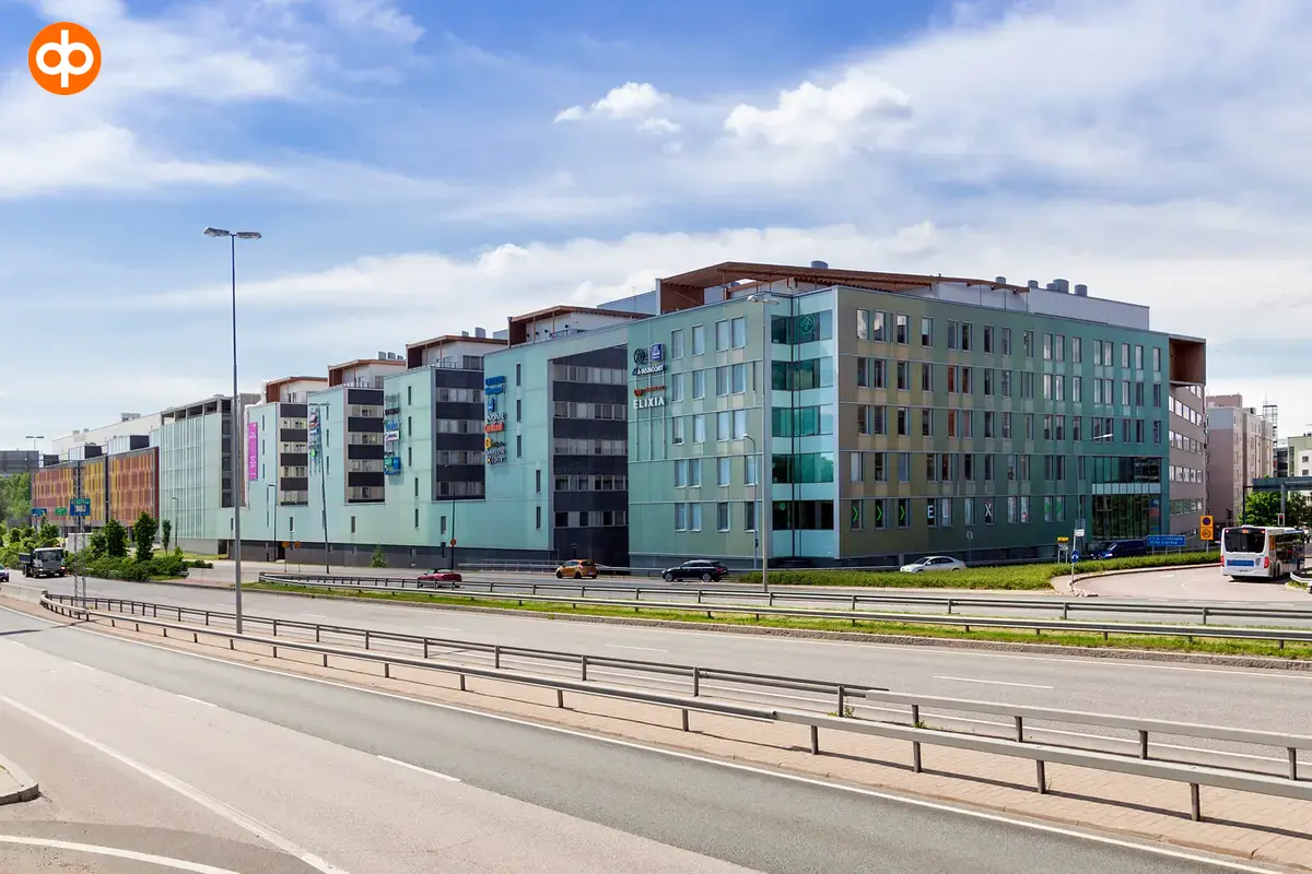 Alberga Business Park – The facade of the D building seen across the road. Gray-green real estate complex. A multi-lane highway in front. Blue sky in the background.