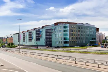 Alberga Business Park – The facade of the D building seen across the road. Gray-green real estate complex. A multi-lane highway in front. Blue sky in the background.